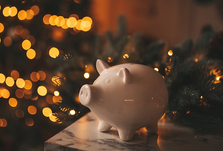 Metas de ahorro Al frente, una alcancía blanca en forma de cerdito sobre una superficie de mármol; detrás, luces cálidas y un árbol de Navidad desenfocado evocan ahorro y metas financieras navideñas para 2025.