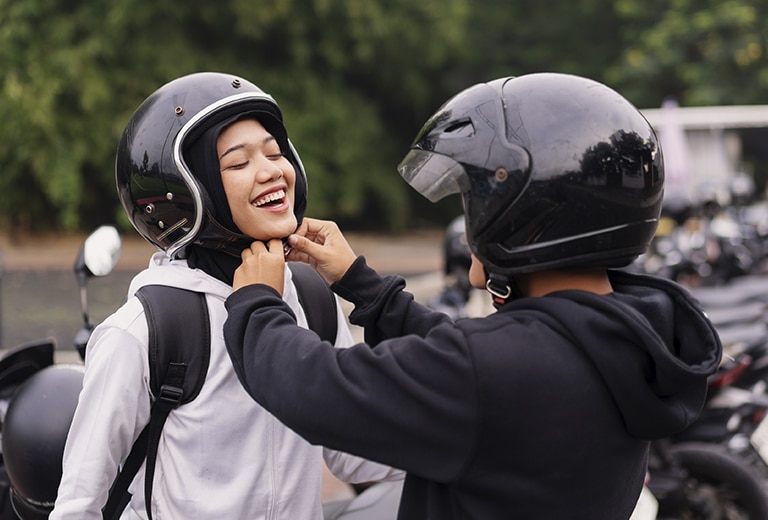 Hacia una movilidad que proteja la vida Una persona con casco negro ayuda a otra a abrochar su casco abierto negro antes de montar en moto, ambas sonriendo en un estacionamiento con más motocicletas al fondo, representando el uso responsable del casco como medida de seguridad vial.