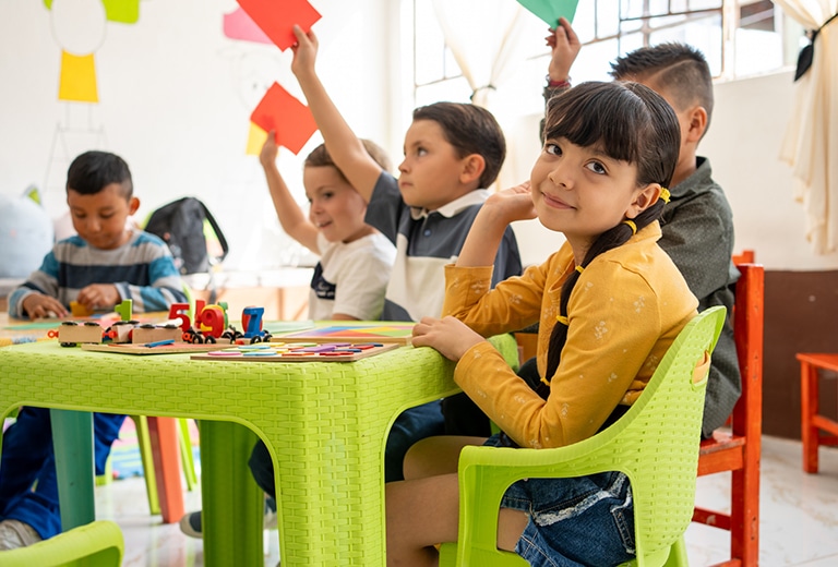 Reducir la pobreza sí es posible Niña de primaria sentada en una silla verde en un aula, sonriendo a cámara mientras otros niños levantan tarjetas de colores y juegan con materiales educativos sobre una mesa, simbolizando aprendizaje y oportunidades.