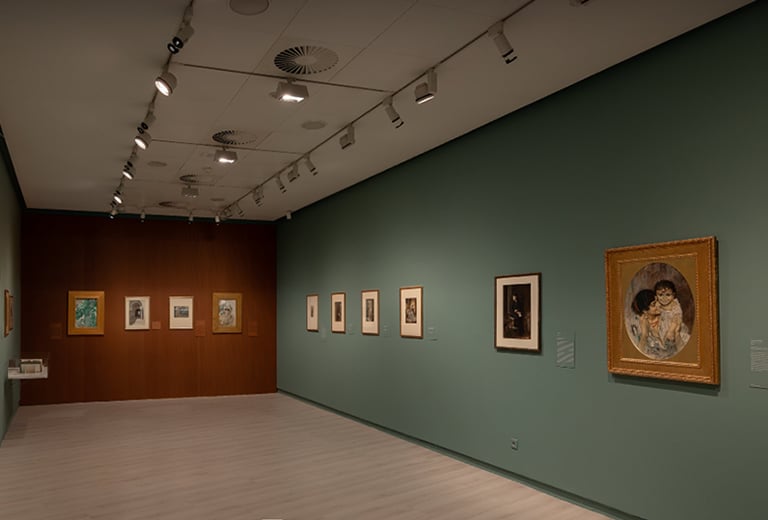 Día Internacional de los Museos Sala de museo con paredes verdes y marrones, varias obras enmarcadas expuestas a ambos lados, iluminación de galería y un cuadro destacado en primer plano.